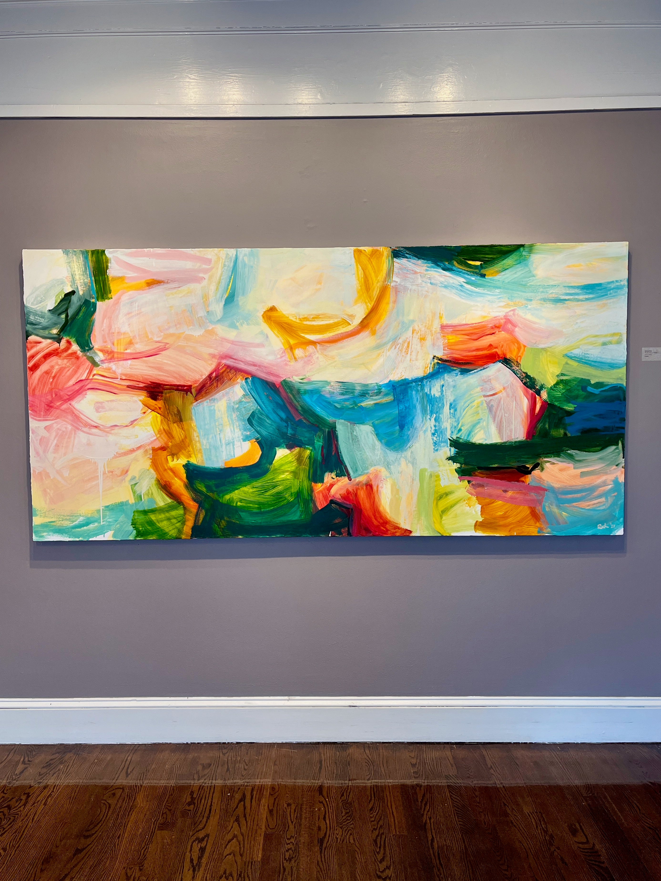An abstract canvas painting with expressive brushstrokes in blue, green, pink, yellow, orange, and white, displayed on a gray wall above a dark wood floor.