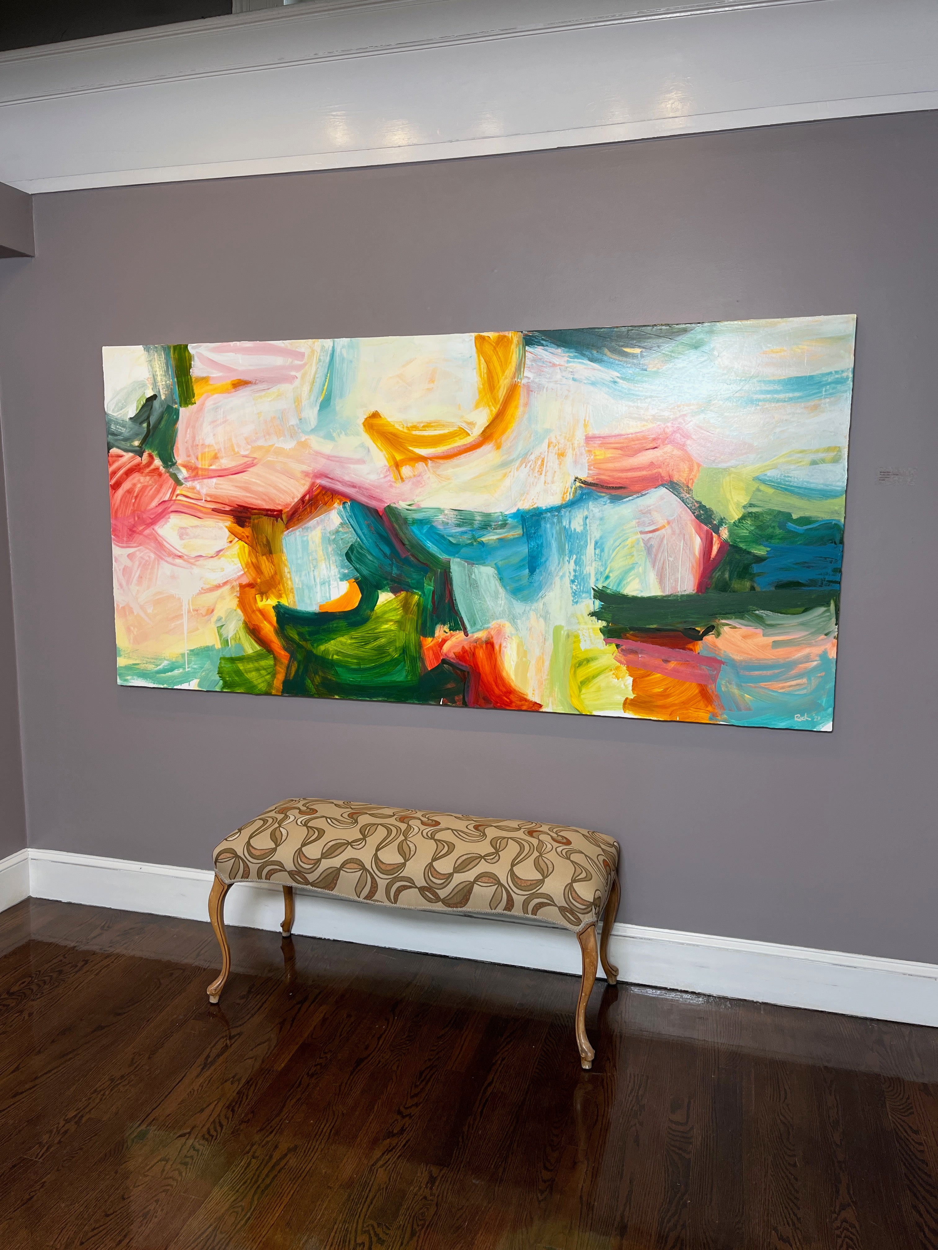 A large rectangular abstract canvas painting with vibrant brushstrokes in pink, blue, green, yellow, and white, displayed on a gray wall above a patterned upholstered bench with wooden legs.