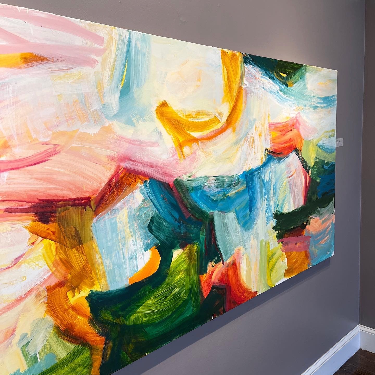 A large abstract canvas painting with expressive brushstrokes in pink, blue, green, yellow, orange, and white, displayed on a dark gray wall in an interior room.