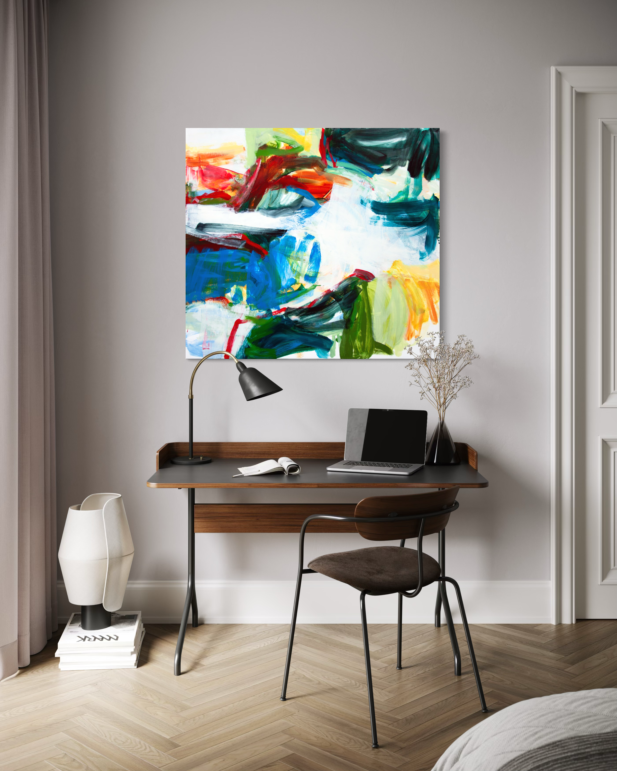 A colorful abstract canvas print with bold brushstrokes of blue, green, red, yellow, and white, displayed on a light gray wall above a wooden desk with a laptop, lamp, and chair.