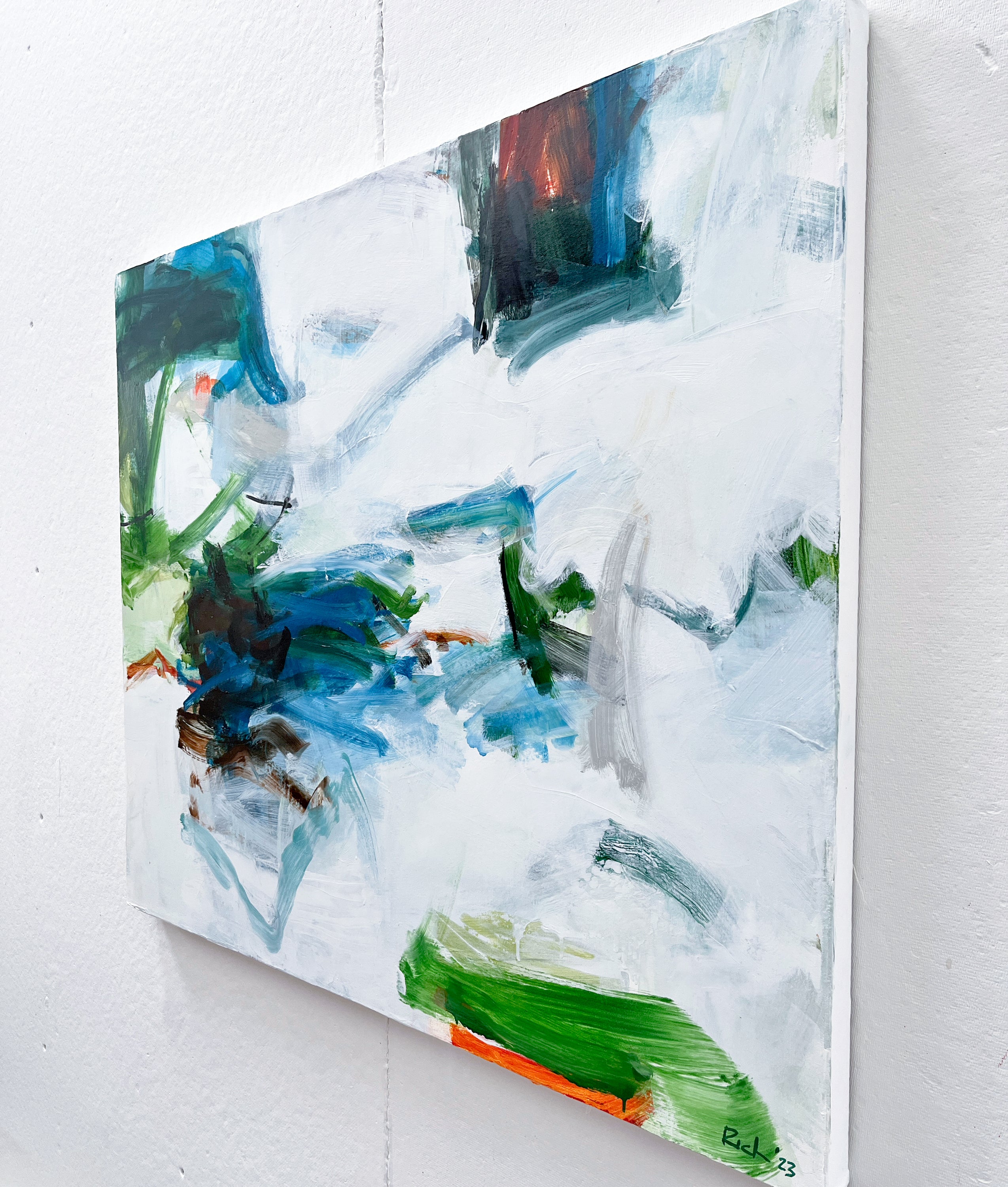 An abstract canvas painting with expressive brushstrokes in white, blue, green, gray, black, and touches of orange, shown hanging on a white wall.