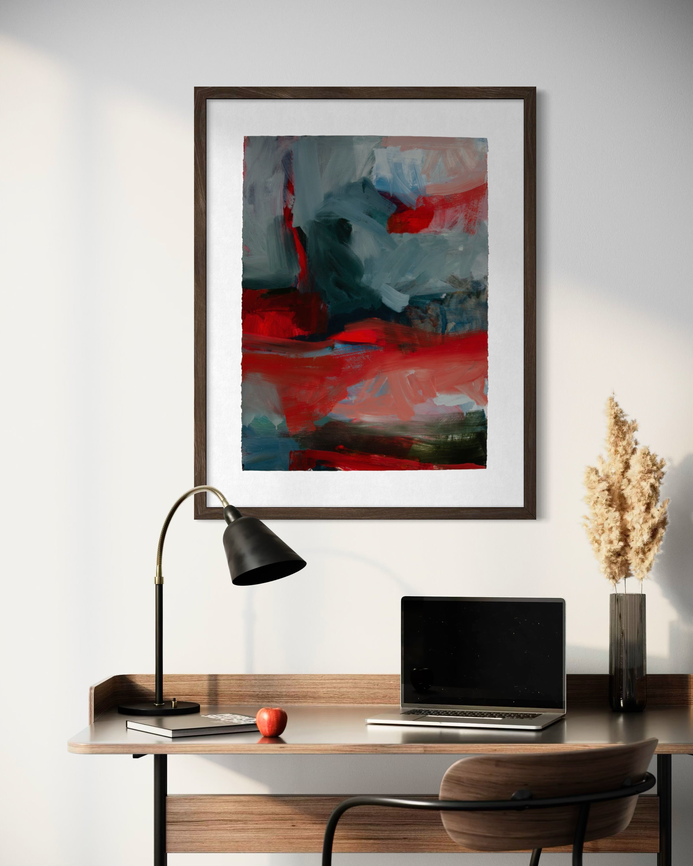 A framed abstract art print featuring bold red, gray, and blue brushstrokes, displayed on a white wall above a wooden desk with a laptop, lamp, and decorative vase.
