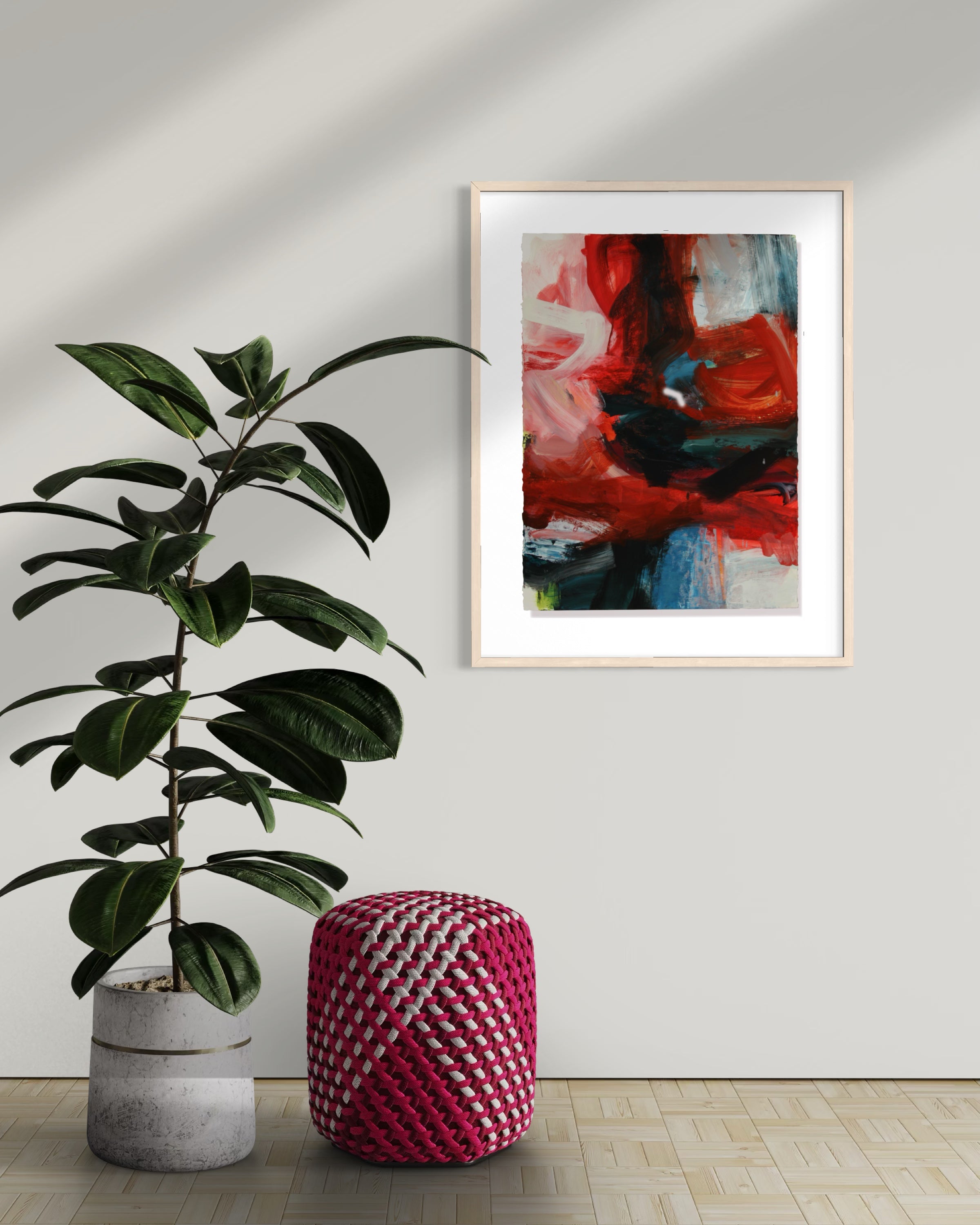 A framed abstract painting with bold red, pink, black, and blue brushstrokes hangs on a light gray wall in a styled room setting, next to a potted green plant and a pink and white woven pouf.