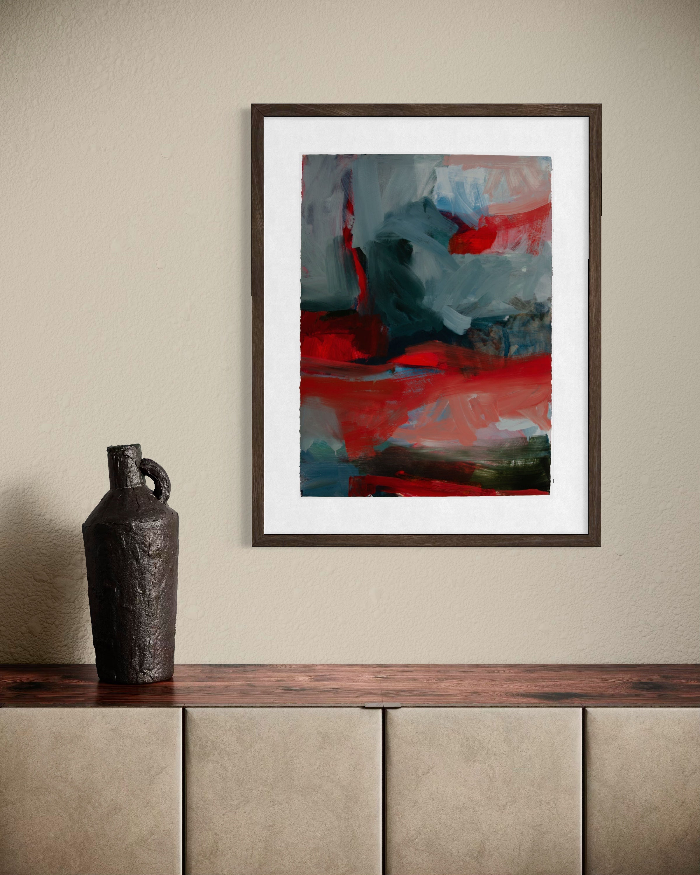 A framed abstract art print featuring bold red, gray, and blue brushstrokes, displayed on a beige wall above a wooden console table with a dark textured vase.