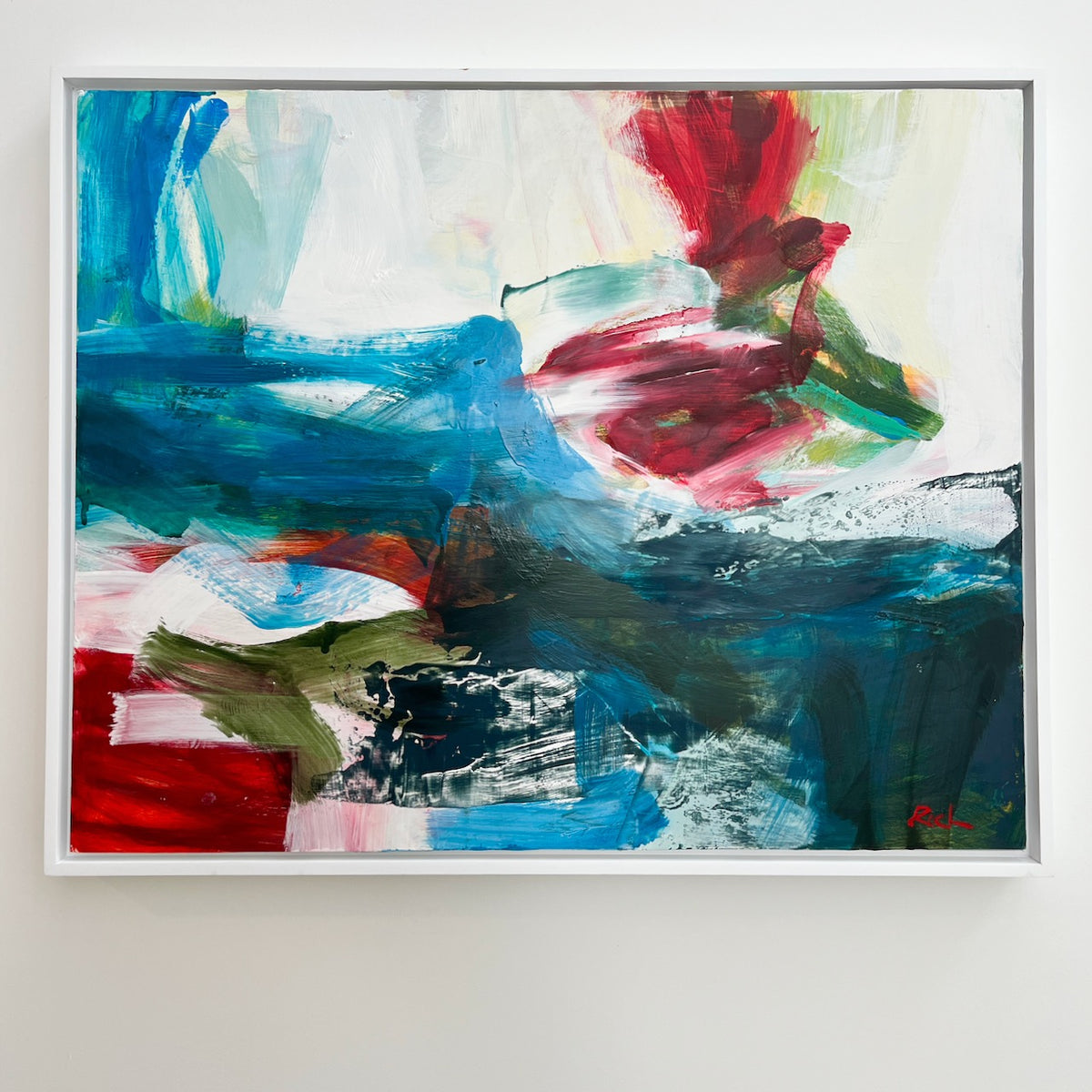 An abstract framed painting by Michael Rich featuring expressive brushstrokes in blue, red, green, white, and black, displayed on a light gray wall.