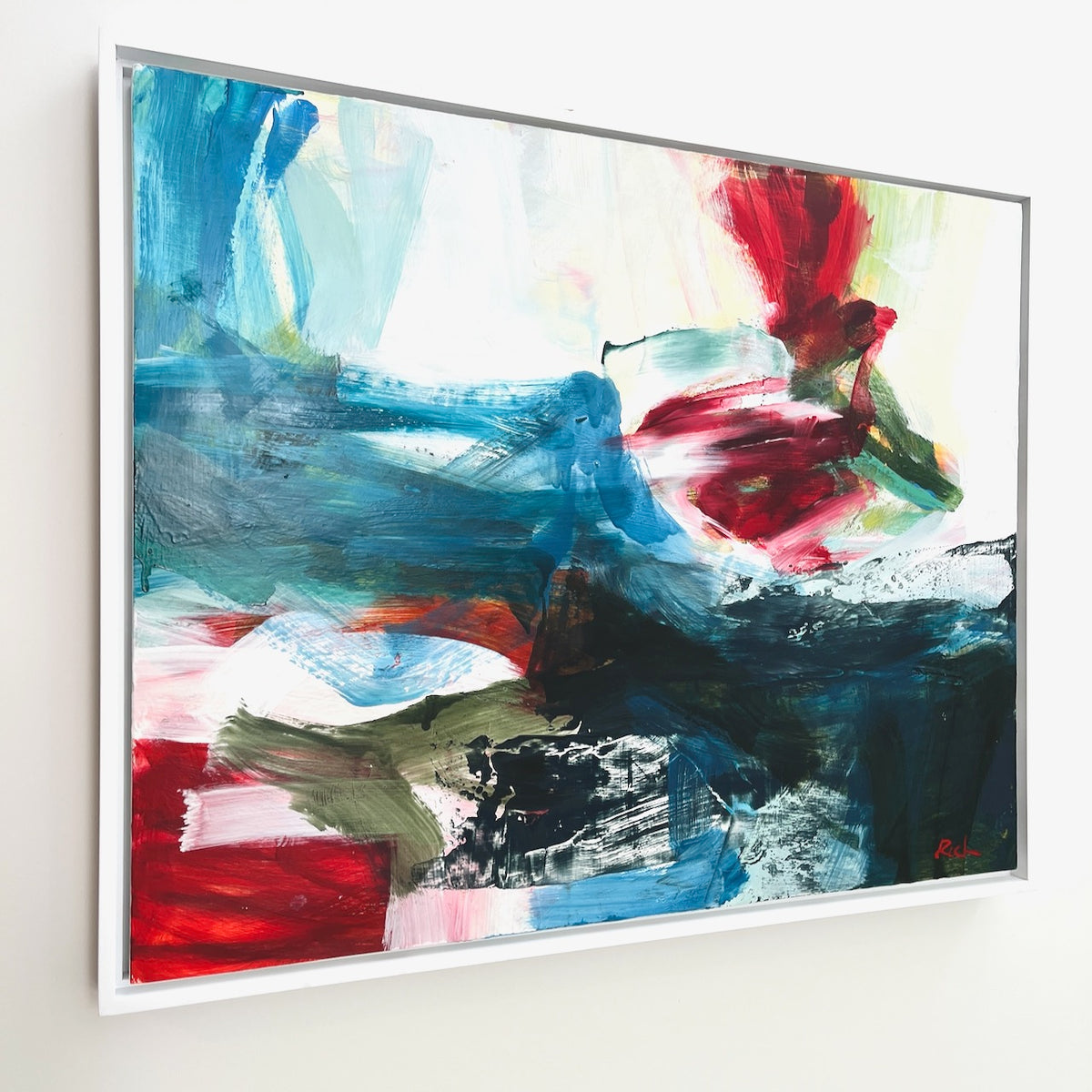An abstract oil on panel painting by Michael Rich with expressive brushstrokes in blue, red, white, black, and green, presented in a white frame and hanging on a light wall.