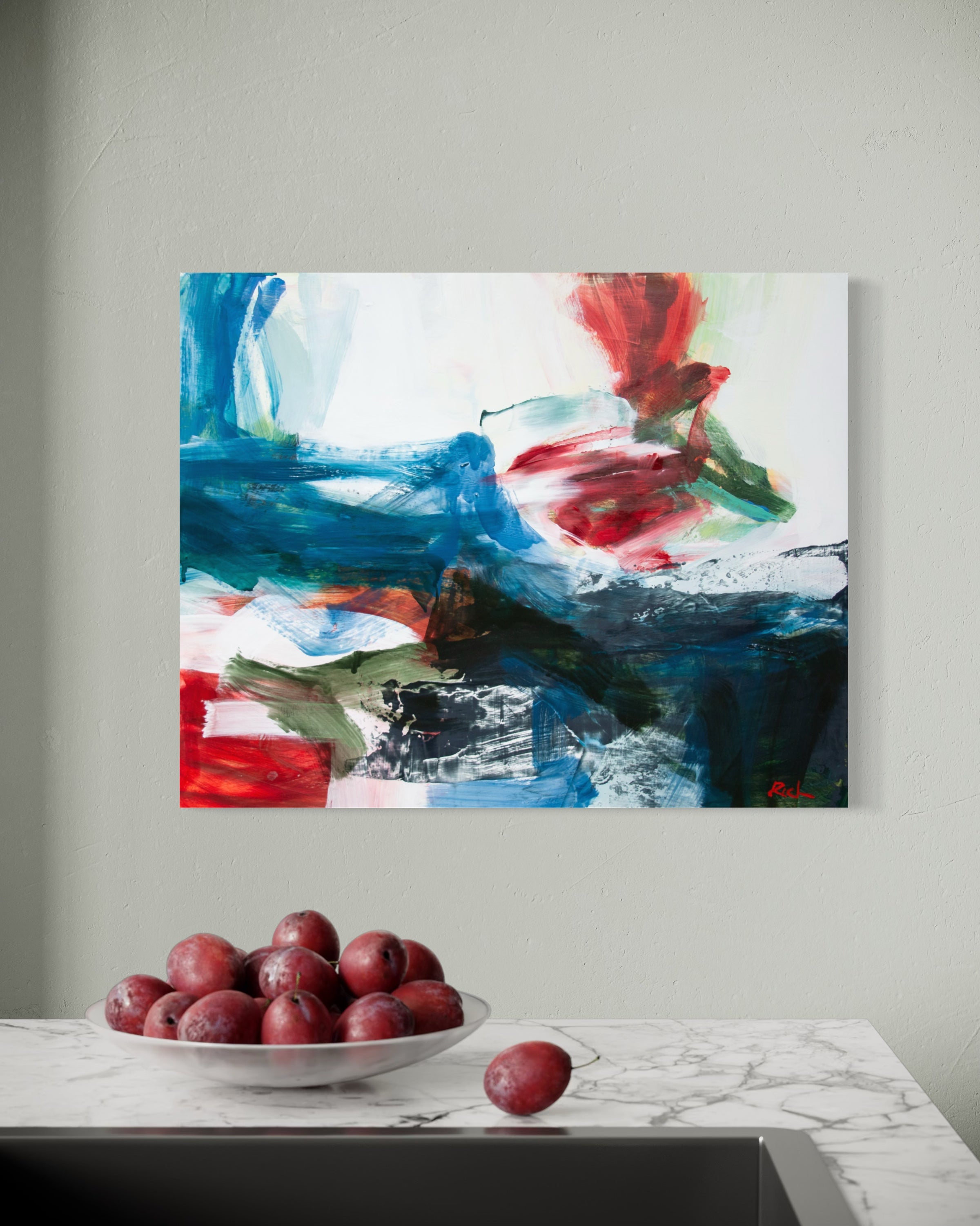 Abstract canvas wall art featuring bold blue, red, green, black, and white brushstrokes, displayed on a light gray wall above a marble countertop with a bowl of red plums.
